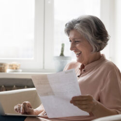 Cheerful senior retired woman paying bills on Internet, counting budget, using online app on laptop at home, typing, holding paper document, invoice, receipt, smiling, laughing